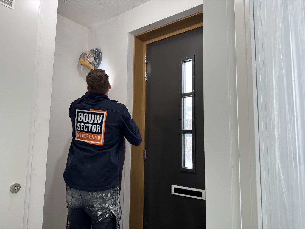 Vakman van Bouwsector Nederland schuurt met een schuurmachine het plafond en de wand boven een voordeur