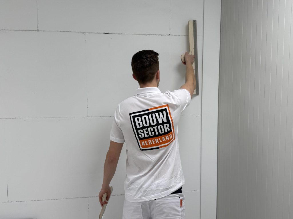 Stukadoor met het Bouwsector-logo op zijn rug strijkt met een lange spaan over een muur van gipsplaat