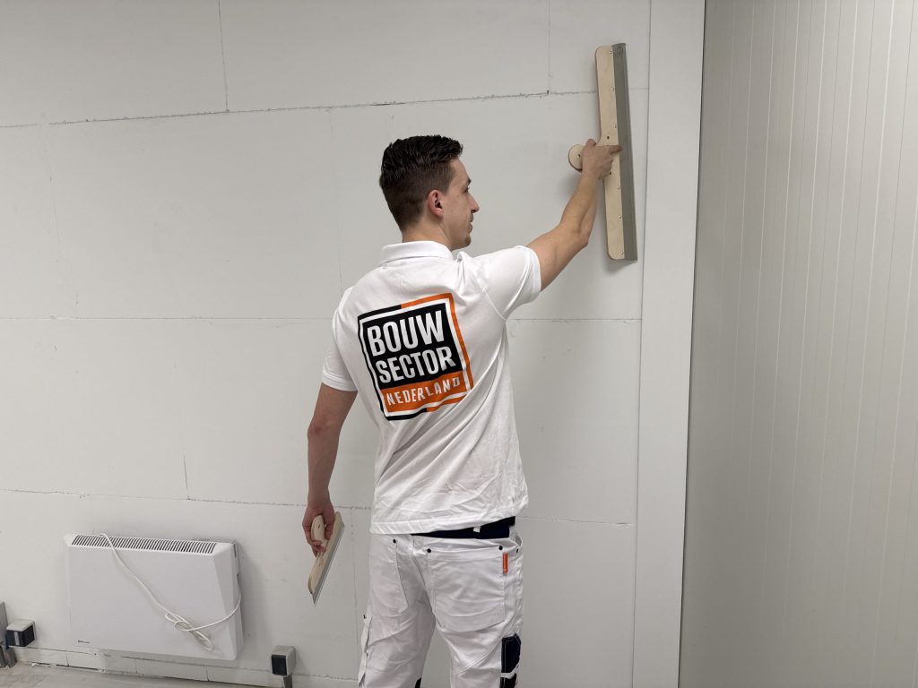 Stukadoor houdt een brede spaan tegen de wand, met een plakspaan in zijn andere hand