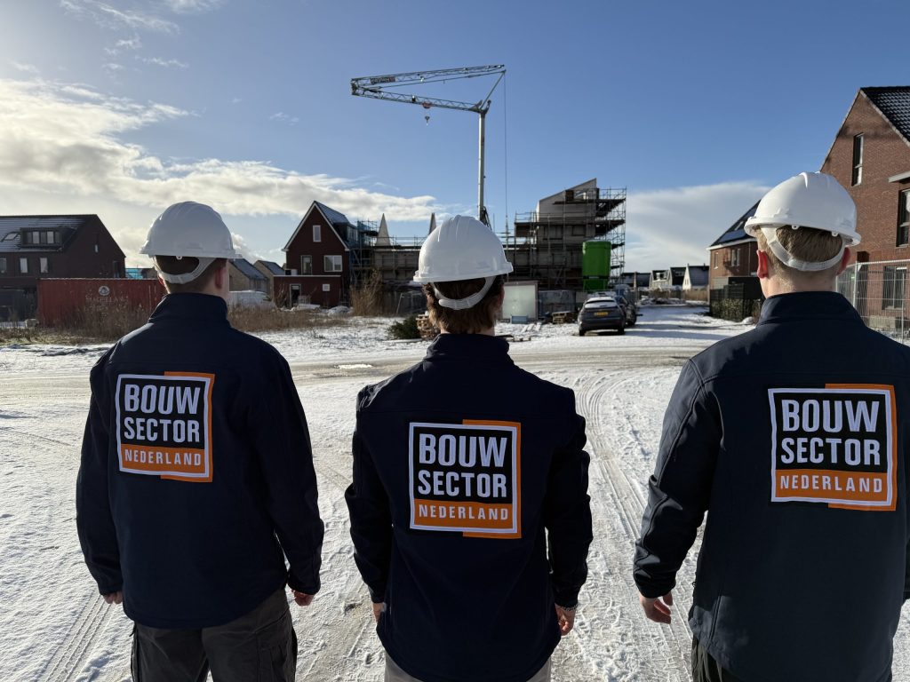Drie vakmensen van Bouwsector Nederland met witte helm op een besneeuwde straat met woningen in aanbouw