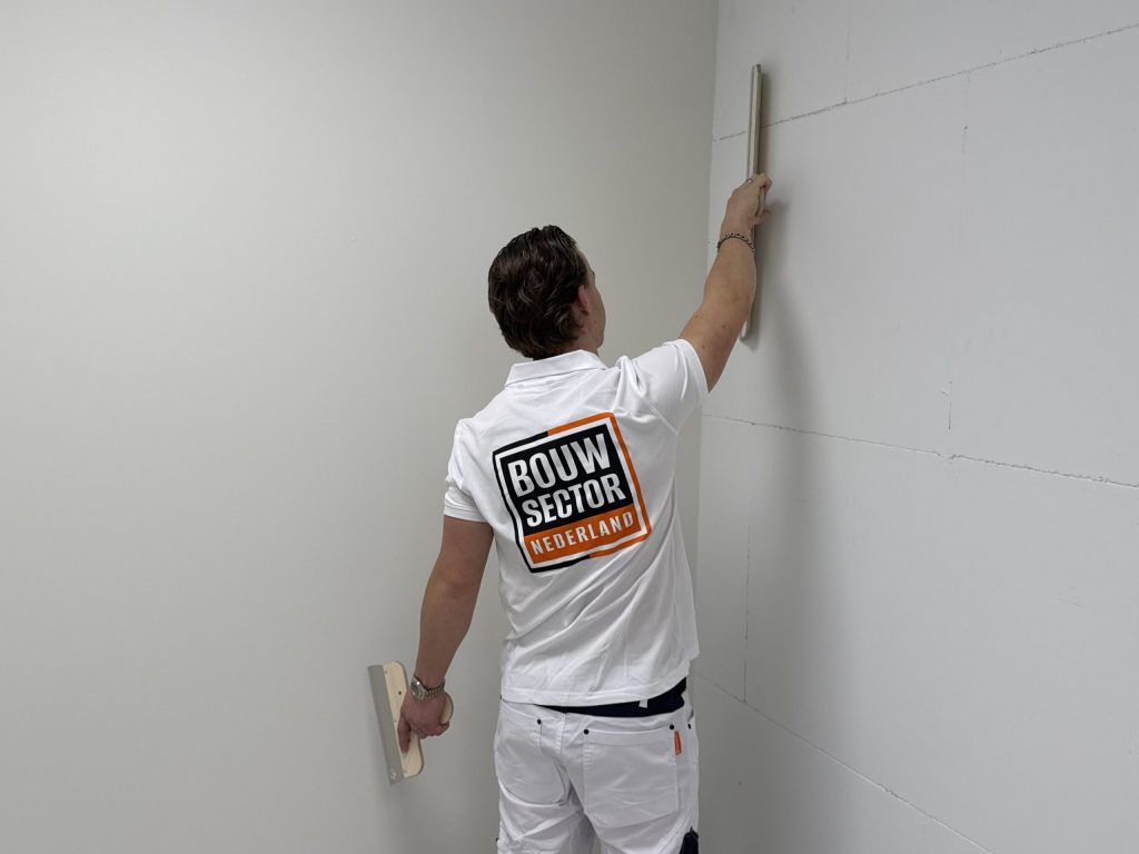 Stukadoor met Bouwsector-logo houdt een lange spaan rechtop tegen de wand vlak bij de hoek